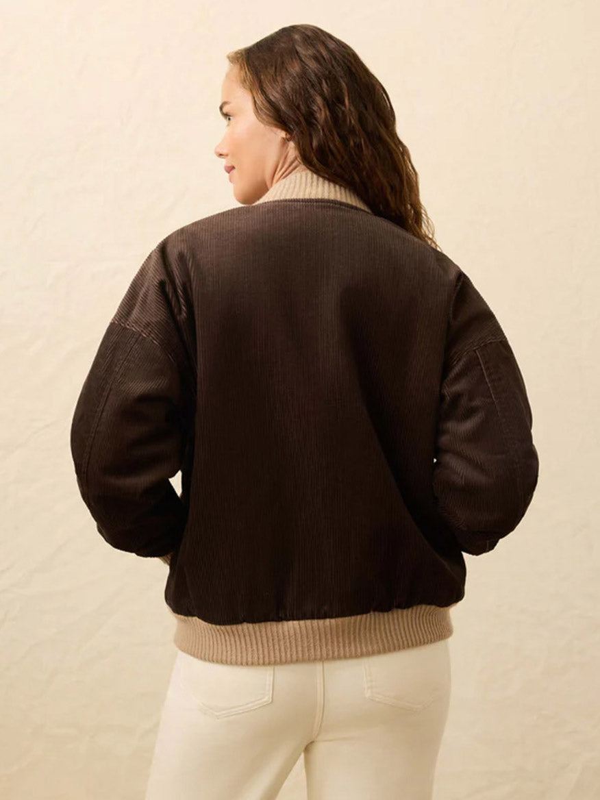 Woman with long brown hair wears the Faherty Brand Heritage Cord Jacket in chocolate brown, featuring a beige collar and cuffs, paired with white pants, shown from the back against a plain background.