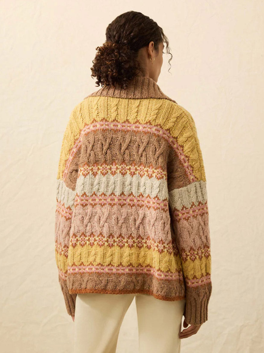 A person with curly hair stands facing away, wearing the Faherty Brand Highland Fairisle Cardigan in Lakeside Fairisle Haze, featuring yellow, brown, white, and pink patterns, paired with cream-colored pants.