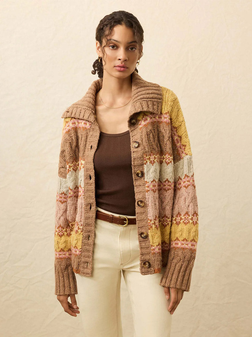 A woman in a brown tank, cream pants, and the Faherty Brand Highland Fairisle Cardigan in Lakeside Fairisle Haze, crafted from soft Merino wool in brown, pink, yellow, and cream, stands against a neutral background.