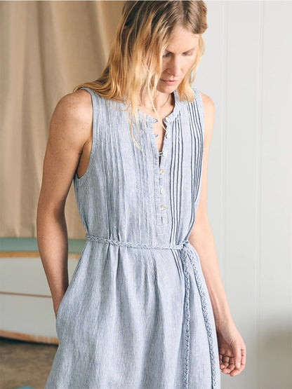 A woman in a sleeveless, striped, button-up Faherty Brand Isha Midi Dress in Blue Mini Stripe with a waist tie, standing in a room with a soft focus background.