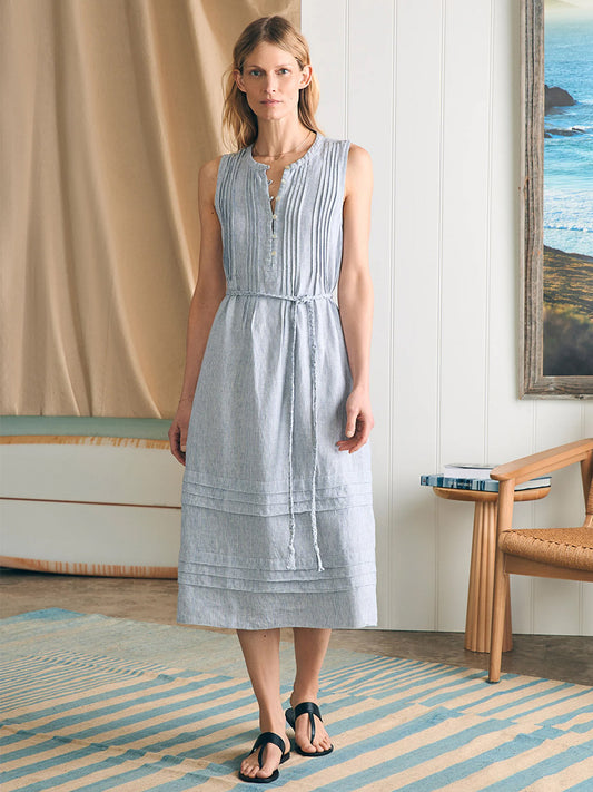 A woman standing in a room with a sea view, wearing the Faherty Brand Isha Midi Dress in Blue Mini Stripe and black sandals.
