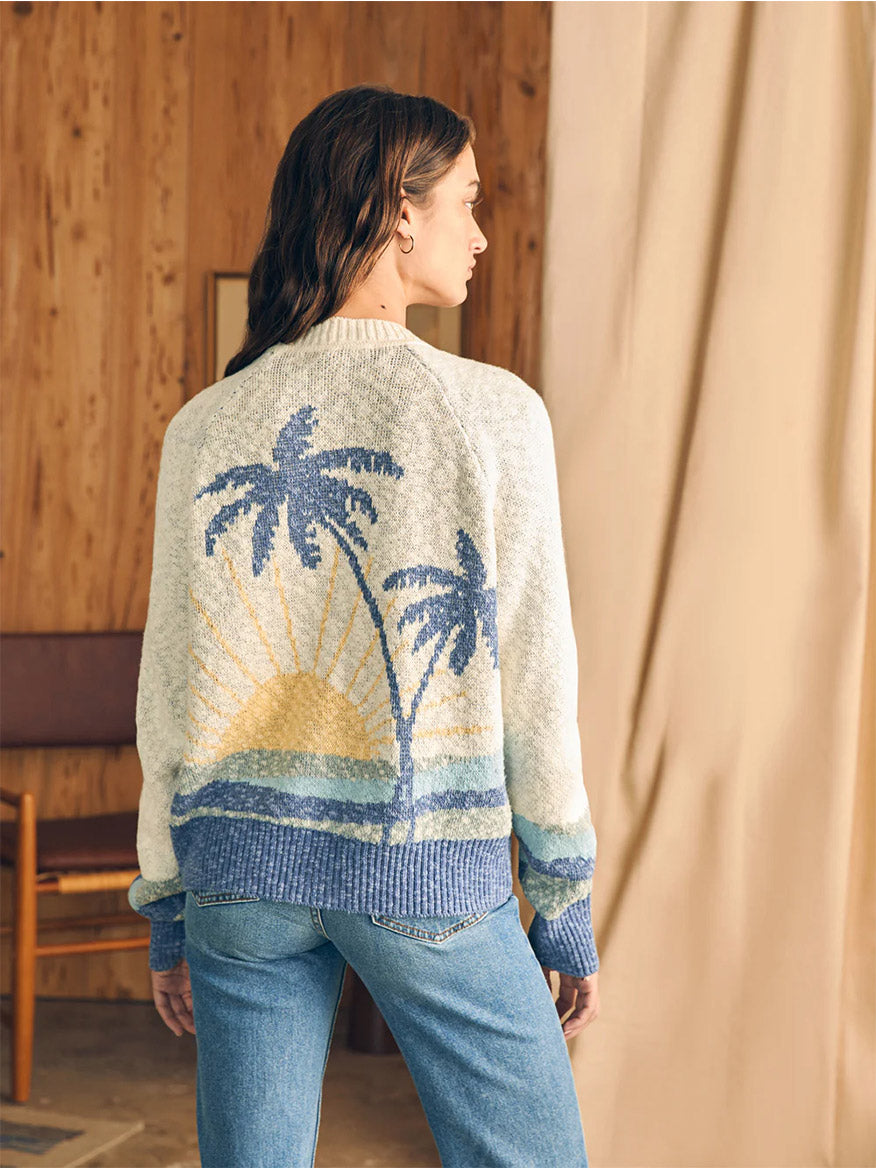 Woman viewed from behind wearing an organic cotton Faherty Brand Island Time Cardigan in Coastal Capri with a palm tree and sunset design, standing in a wood-paneled room.