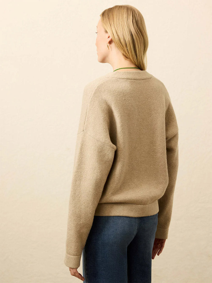 A woman with blonde hair, wearing the Faherty Brand Jackson V-Neck Popover in Oatmeal Heather and blue jeans, stands facing away from the camera against a plain background.