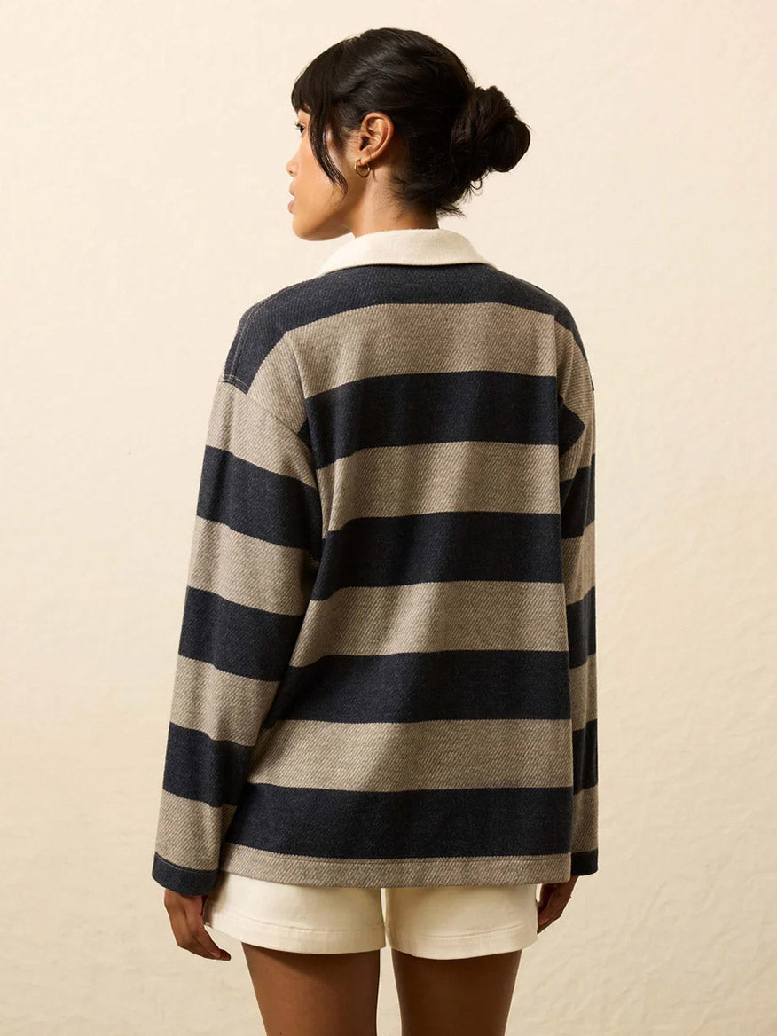 A person with dark hair in a bun stands with their back to the camera, wearing white shorts and the Faherty Brand Legend™ Sweater Rugby Popover in Navy Rugby—a long-sleeve, navy and beige classic striped shirt.