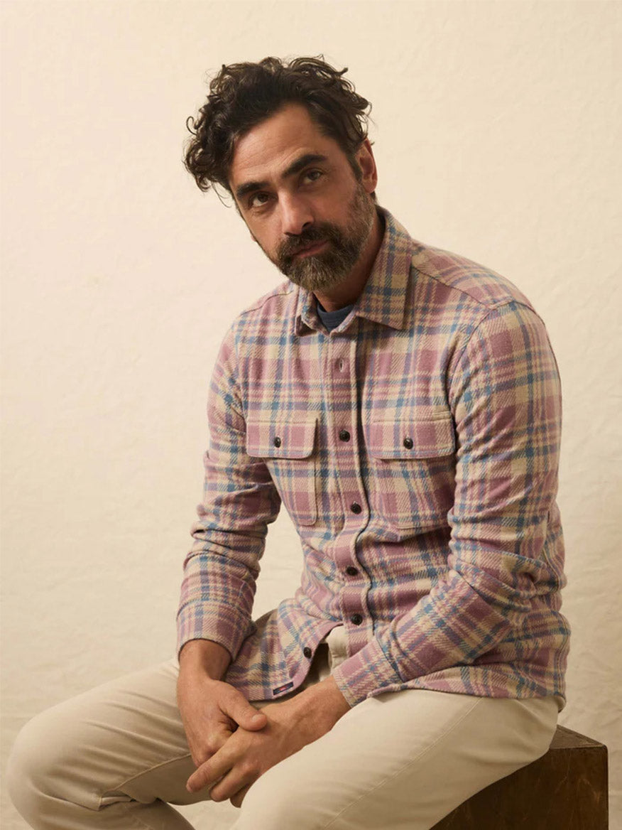 A man with dark curly hair and beard wears the Faherty Brand Legend Sweater Shirt in Cedar Shore Plaid and light pants, seated on a stool against a plain light background.