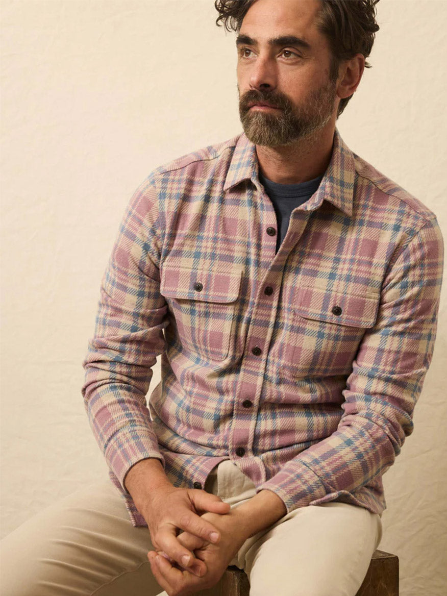 A bearded man sits on a wooden stool, wearing the Faherty Brand Legend Sweater Shirt in Cedar Shore Plaid with light pants, set against a plain, light background.