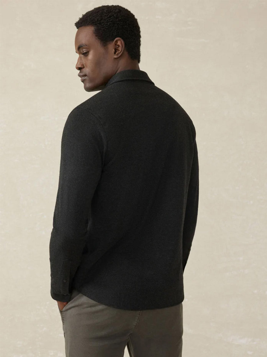 A man faces away, wearing the Faherty Brand Legend Sweater Shirt in Heathered Black Twill and gray pants, set against a light neutral background.