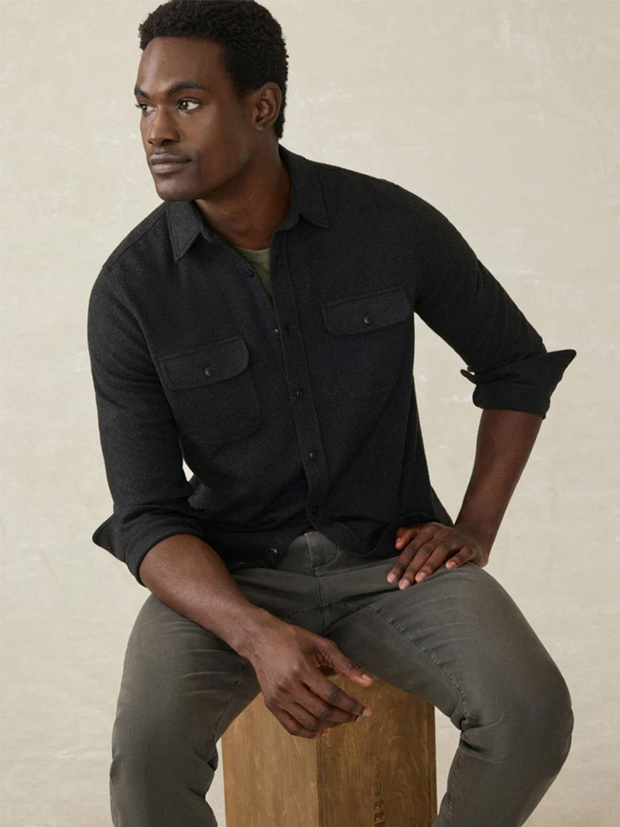 A man in a Faherty Brand Legend Sweater Shirt in Heathered Black Twill and gray pants sits on a wooden block, looking to his left against a neutral background.