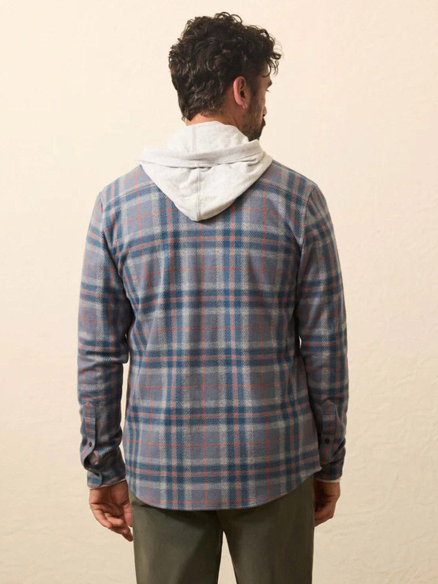 A man faces away from the camera, wearing a gray hoodie under a Faherty Brand Legend Sweater Shirt in Three Lakes Plaid and green pants, set against a plain beige background.