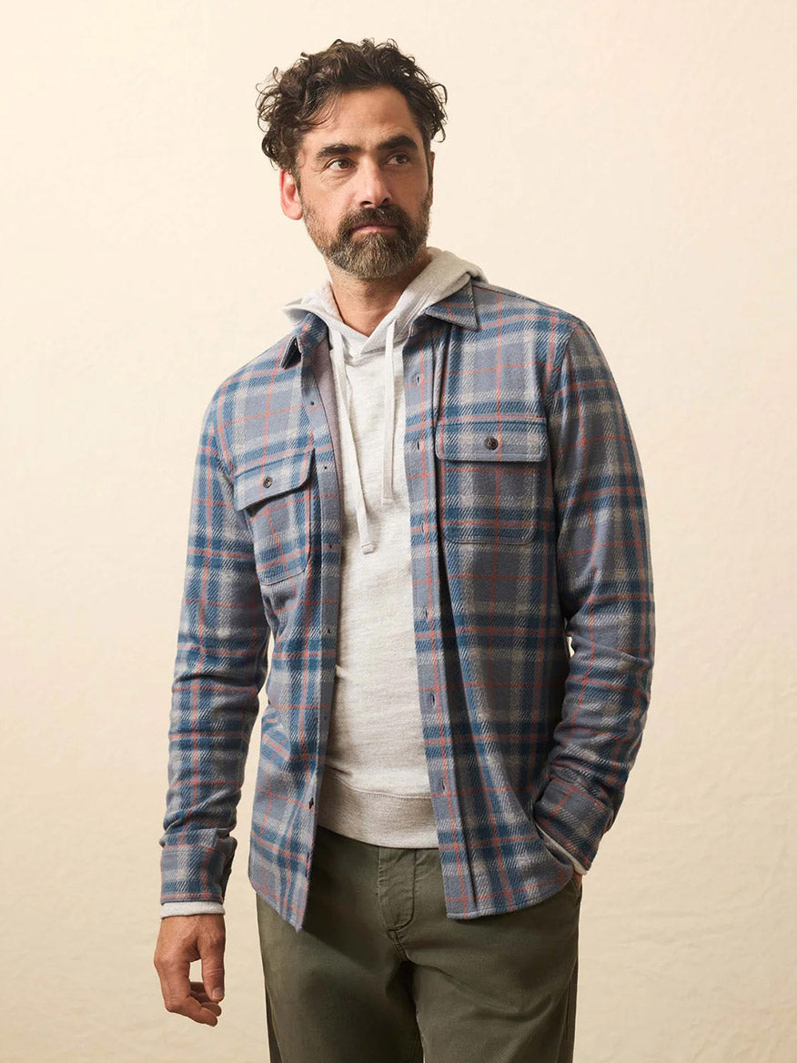 A man with curly dark hair and a beard is wearing a Faherty Brand Legend Sweater Shirt in Three Lakes Plaid over a light hoodie and green pants, standing in front of a plain light background.