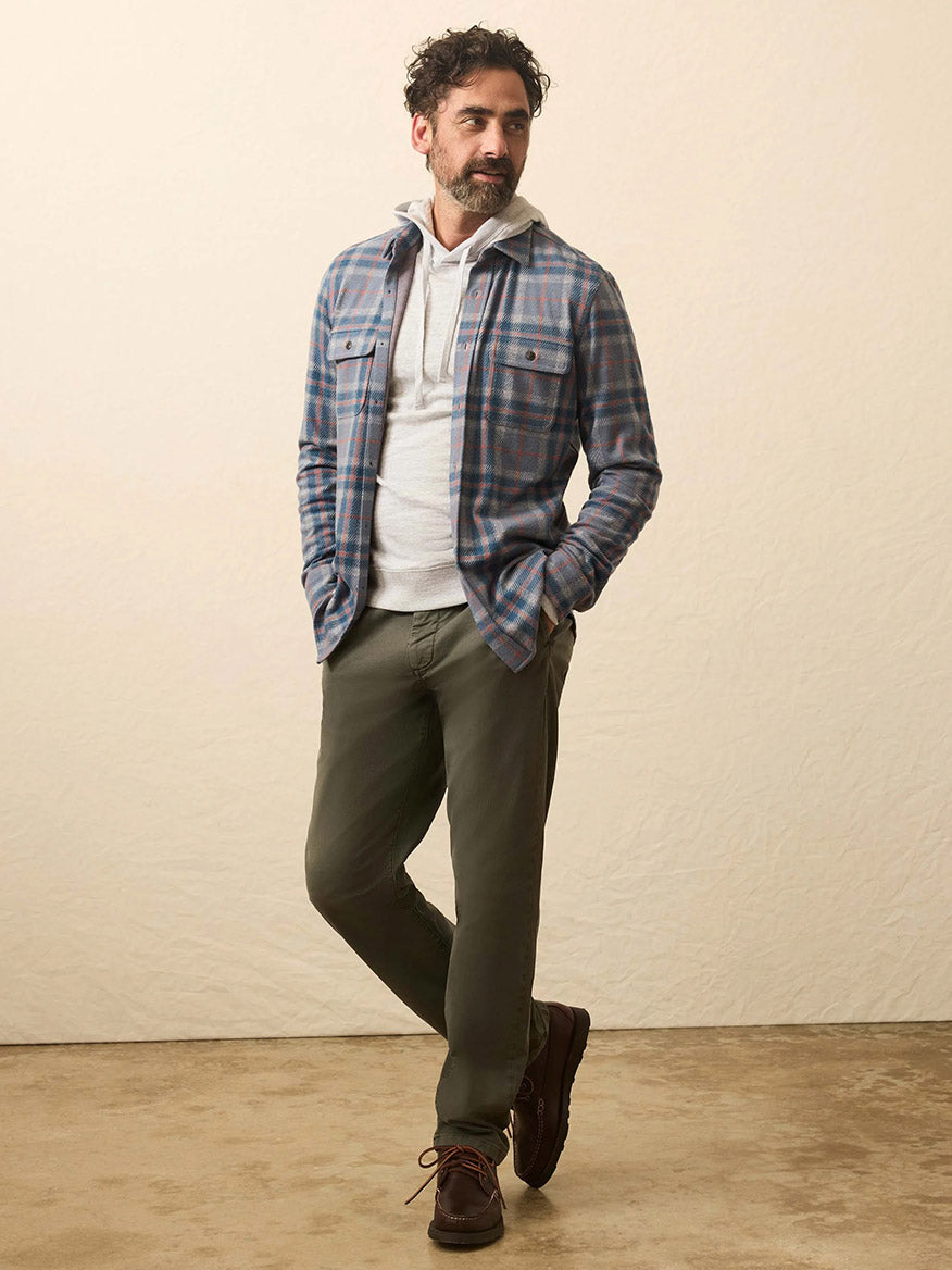 A man stands indoors wearing a gray hoodie under a blue Faherty Brand Legend Sweater Shirt in Three Lakes Plaid, olive green pants, and brown shoes, looking slightly to the side.