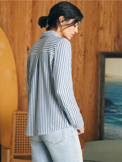 Woman in a Faherty Brand Legend Sweater Shirt in Navy Blazer Stripe and jeans standing in a room with wooden walls and a mirror reflecting a painting of the sea.