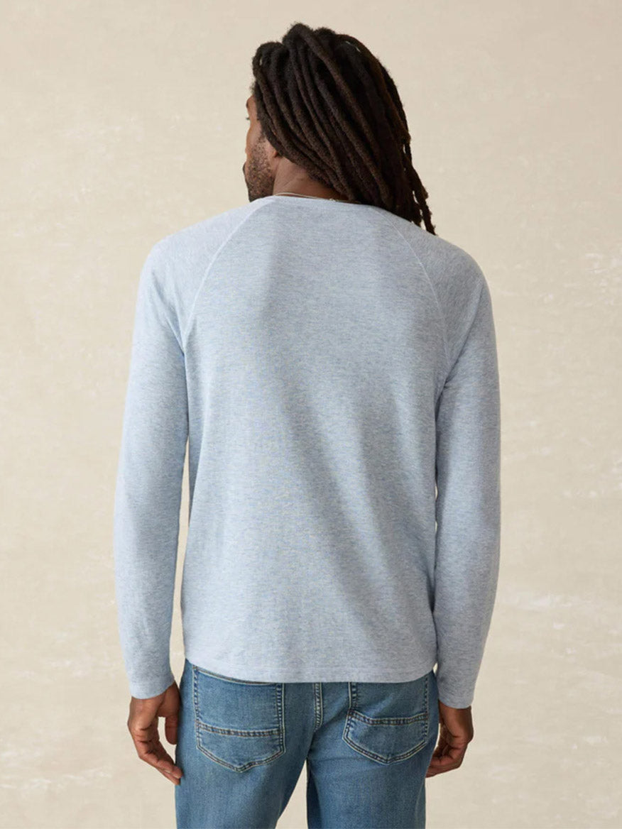 A man with long hair stands facing away, wearing the Faherty Brand Cloud Cotton Long-Sleeve Henley in Light Blue Heather and blue jeans against a plain beige background.