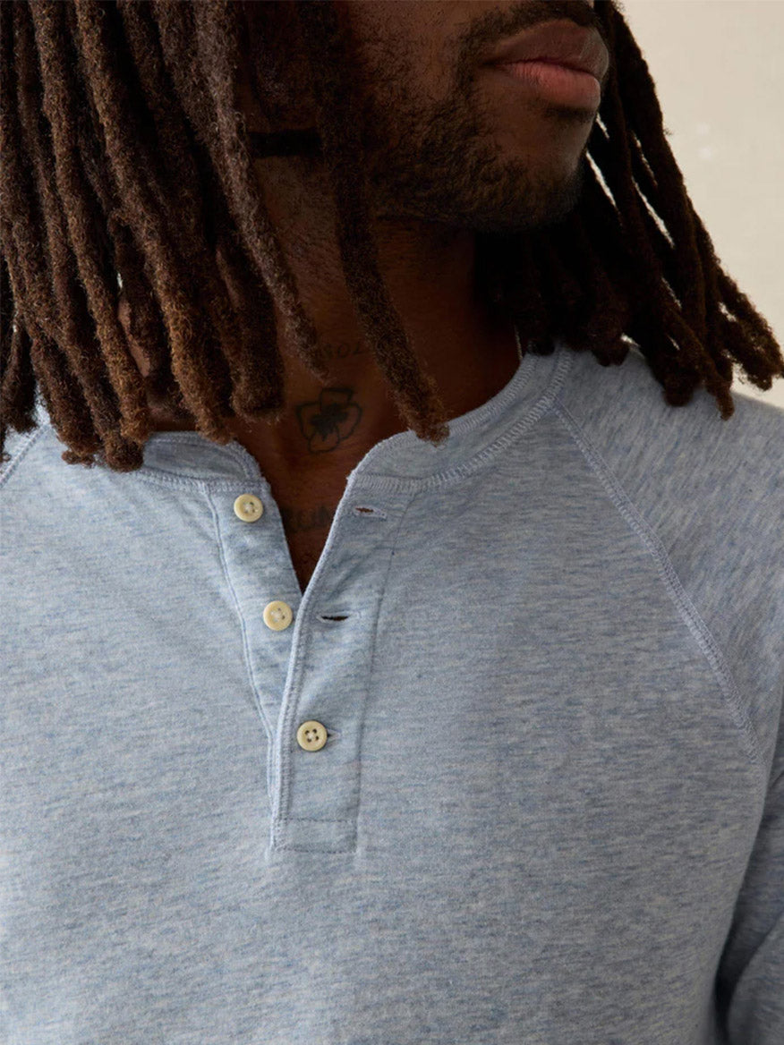 A person with long, brown, textured hair and a neck tattoo wears the Faherty Brand Cloud Cotton Long-Sleeve Henley in Light Blue Heather, its double-knit Pima cotton base layer highlighted by three fastened buttons.