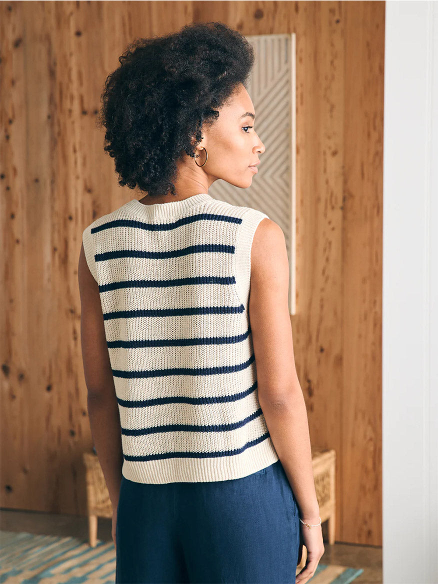 A woman with curly hair stands in a room, wearing a Faherty Brand Miramar Linen Muscle Tank in Montauk Stripe and navy pants, facing away and looking to the side.