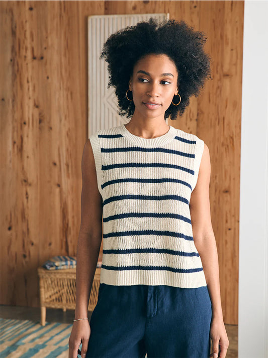 A young woman wearing a Faherty Brand Miramar Linen Muscle Tank in Montauk Stripe and navy pants stands in a room with wooden walls, looking towards the camera.