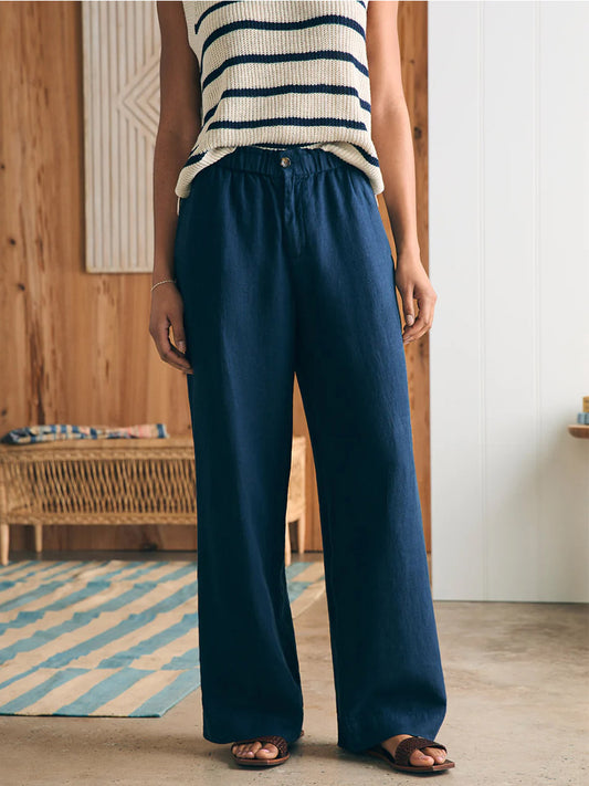 A woman standing in a room, wearing Faherty Brand Monterey Linen Pant in After Midnight and a striped sleeveless top, with one hand in her pocket.