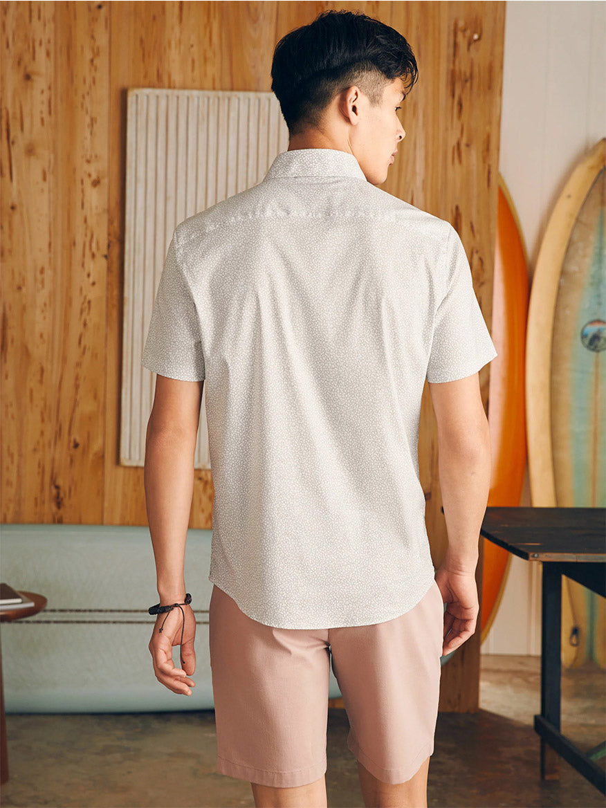 A man stands facing away from the camera in a room with surfboards, wearing a white Faherty Brand Movement Short-Sleeve Shirt in Ivory Cliff Floral and pink shorts.