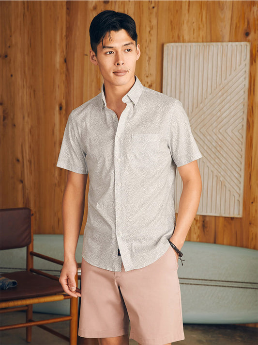 A man in a Faherty Brand Movement Short-Sleeve Shirt in Ivory Cliff Floral and pink shorts stands in a wooden-paneled room, looking to the side.