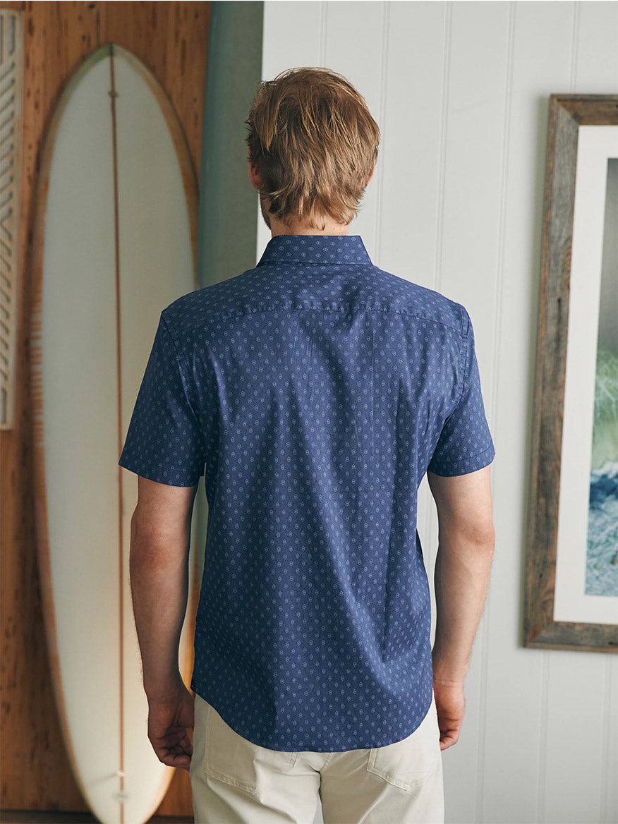 A man viewed from behind wearing a Faherty Brand Movement Short-Sleeve Shirt in Navy Dusk Diamond Print, standing in a room with a surfboard and framed pictures on the wall.