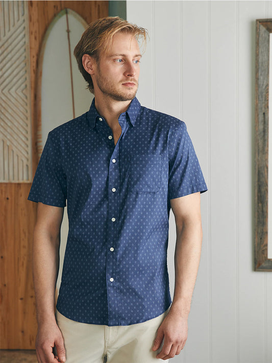 A man in a Faherty Brand Movement Short-Sleeve Shirt in Navy Dusk Diamond Print and beige pants stands in a room with wooden paneling, looking to his left with a thoughtful expression.