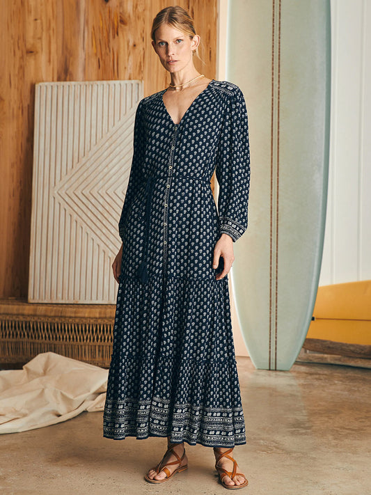 A woman stands in a wooden room wearing the Faherty Brand Orinda Long Sleeve Maxi Dress in Lotus Floral Print and sandals, next to a surfboard leaning against the wall.