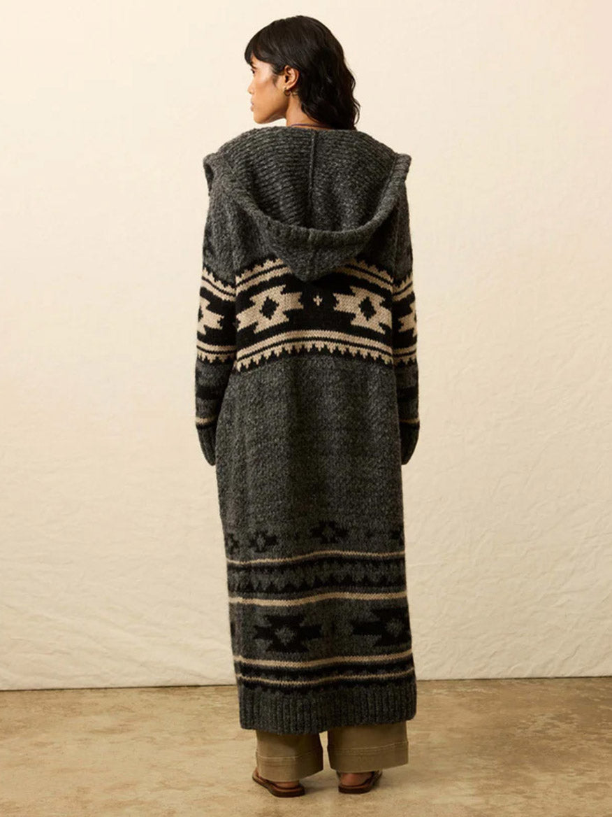 A person stands indoors facing away, wearing the Faherty Brand Steven Paul Judd Chahtah Hooded Duster in Charcoal Chahtah with cream and black geometric patterns, layered over brown pants and sandals.
