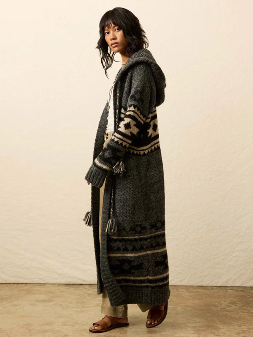 A person stands sideways against a plain background wearing the Faherty Brand Steven Paul Judd Chahtah Hooded Duster in Charcoal Chahtah, paired with light pants and sandals.