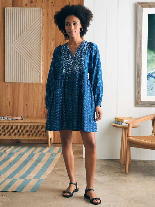 A woman in a Faherty Brand Silk Blend Solstice Mini Dress in Sunburst Mix Print stands in a stylish room with wooden furniture and a striped rug.