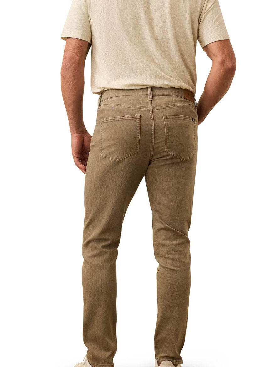 A man stands facing away, wearing a beige short-sleeve shirt and Faherty Brand Stretch Terry 5-Pocket Pant in Dark Rye Heather, shown from the waist down with visible back pockets.