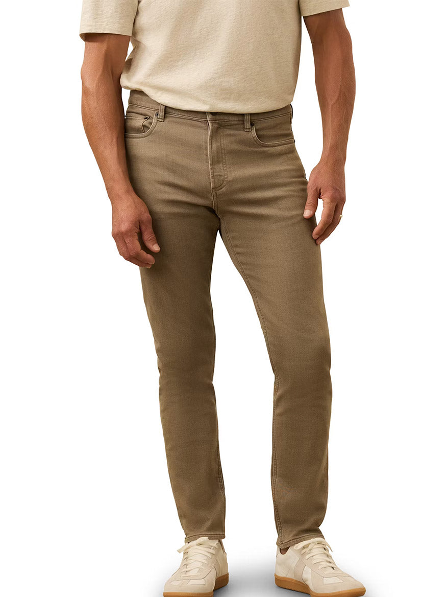 A man wearing a beige t-shirt, Faherty Brand Stretch Terry 5-Pocket Pant in Dark Rye Heather, and white sneakers stands against a plain background.