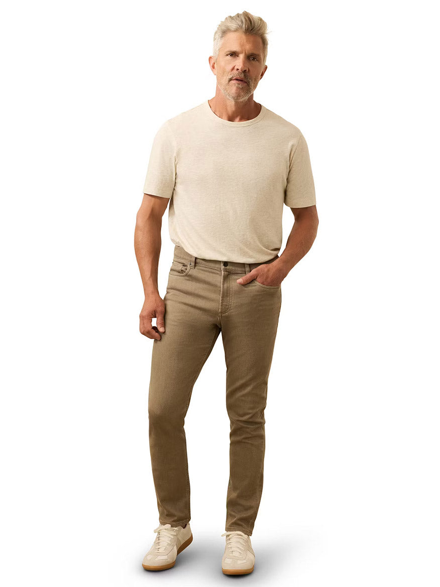 A man with gray hair and a beard stands against a white background, wearing a beige t-shirt, Faherty Brand Stretch Terry 5-Pocket Pant in Dark Rye Heather, and white sneakers.