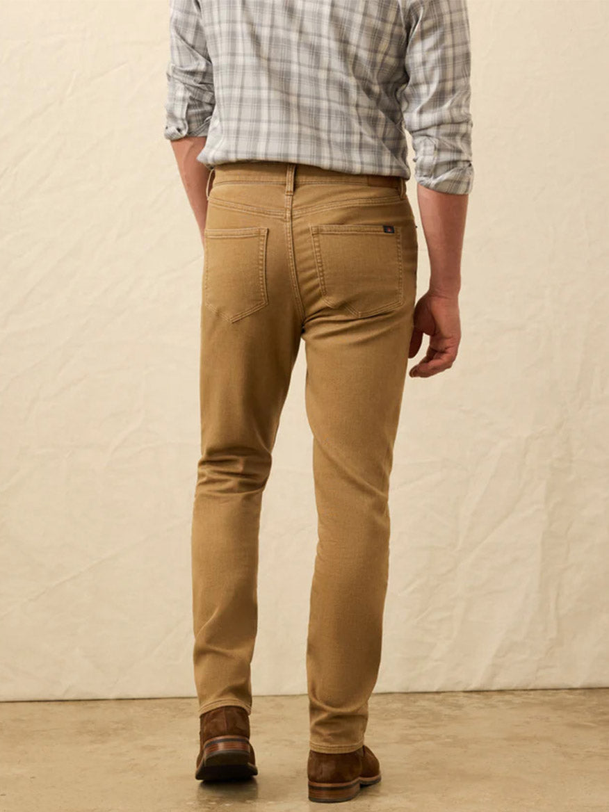 A man faces away in a plaid shirt, brown shoes, and Faherty Brand Stretch Terry 5-Pocket Pant in Elk Brown, highlighting an authentic denim look against a plain light background.