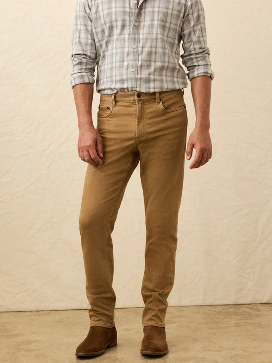 A man stands against a neutral background in a light plaid button-up shirt, Faherty Brand Stretch Terry 5-Pocket Pant in Elk Brown, and brown suede boots for a stylish look.