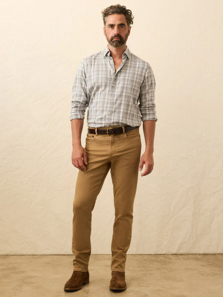 A man with short wavy hair and a beard wears a light gray plaid shirt, Faherty Brand Stretch Terry 5-Pocket Pant in Elk Brown, a brown belt, and brown shoes, standing against a neutral background.