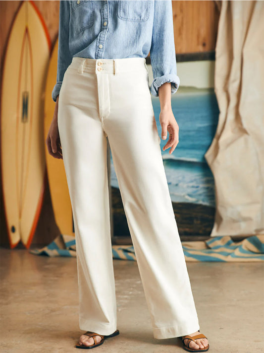 Woman in a casual outfit with a Faherty Brand Stretch Terry Harbor Pant in Egret and white wide-leg pants standing beside a surfboard.