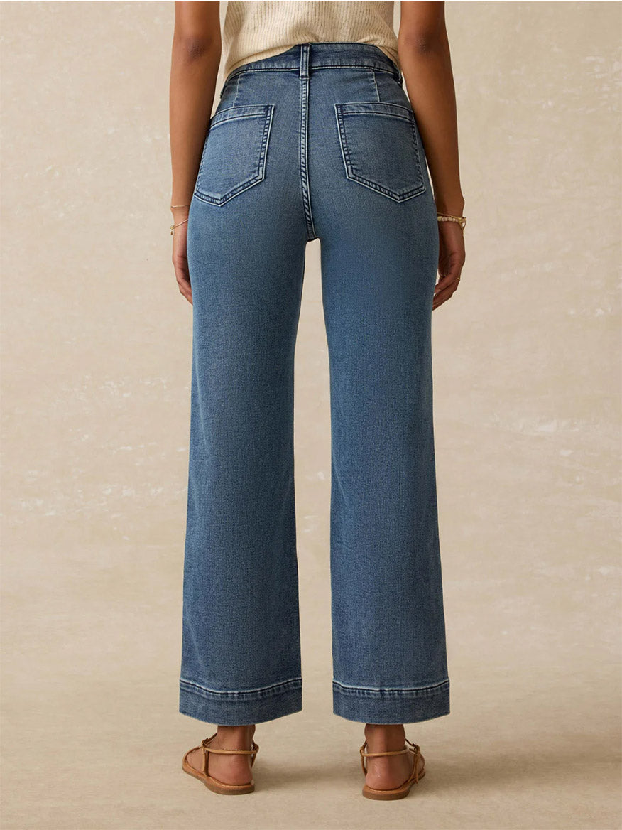 Wearing Faherty Brand Stretch Terry 5-Pocket Ankle Pant in Riverton, a person stands facing away from the camera in relaxed fit blue jeans and sandals against a neutral background.