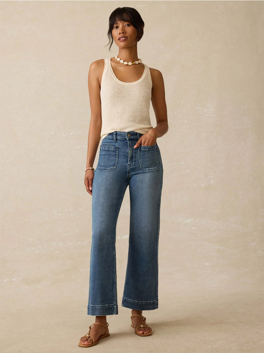 A woman stands against a neutral background wearing a beige crewneck sleeveless knit top, Faherty Brand Stretch Terry 5-Pocket Ankle Pant in Riverton, sandals, and a beaded necklace.