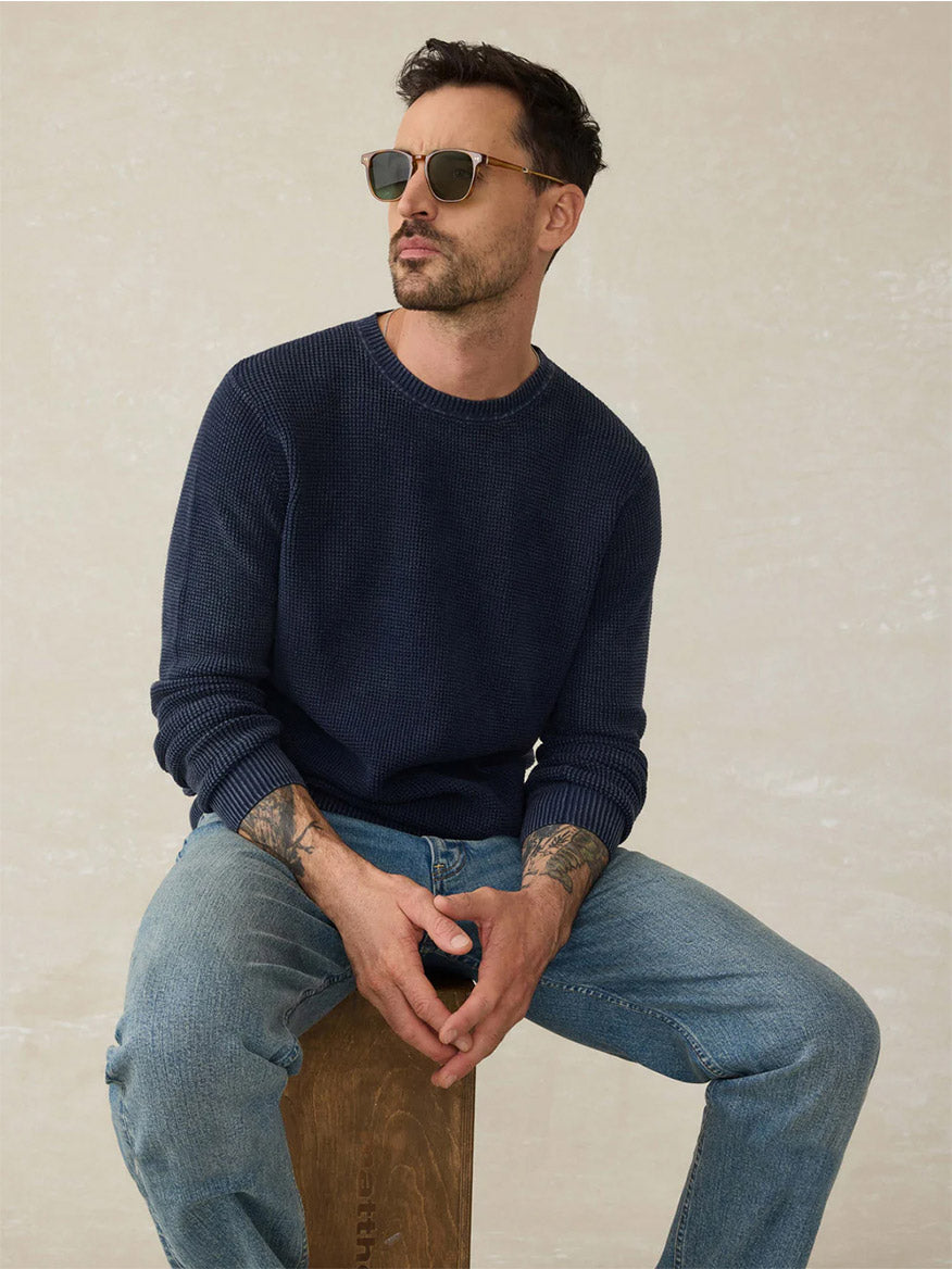 A man with short dark hair and sunglasses sits on a wooden stool, wearing the Faherty Brand Sunwashed Crewneck Sweater in Blue Nights and light blue jeans, against a plain light background.