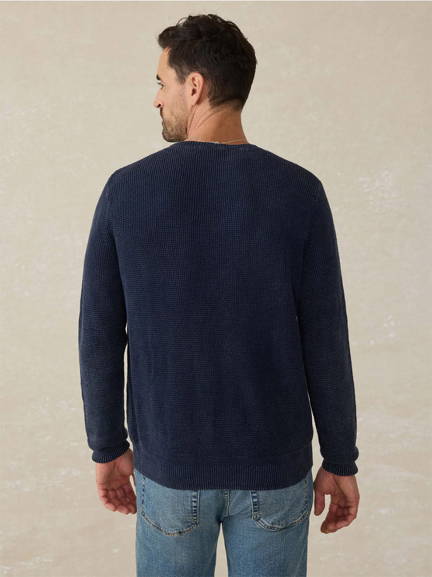 A man stands with his back to the camera, wearing the Faherty Brand Sunwashed Crewneck Sweater in Blue Nights and light blue jeans against a neutral background for a timeless, vintage look.
