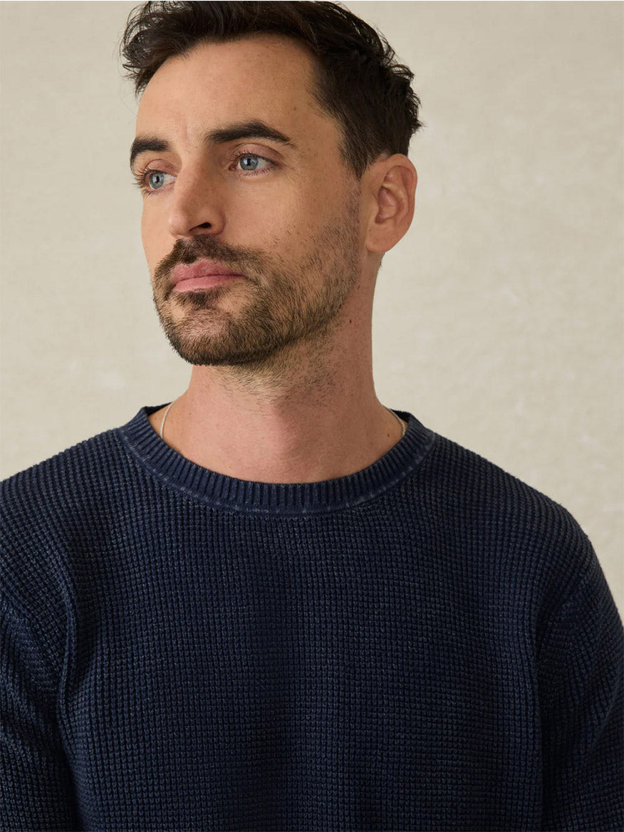 A man with short dark hair, light eyes, and a trimmed beard wears the Faherty Brand Sunwashed Crewneck Sweater in Blue Nights, looking slightly to the side against a neutral background.