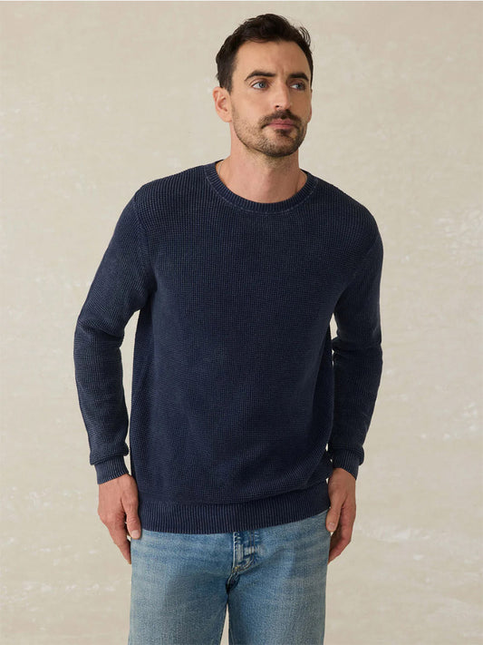 A man with short dark hair and a trimmed beard wears the Faherty Brand Sunwashed Crewneck Sweater in Blue Nights and light blue jeans, standing against a neutral background for a classic, vintage look.