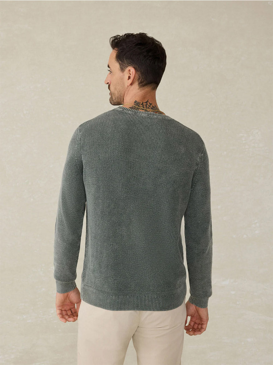 A man stands with his back to the camera in a Faherty Brand Sunwashed Crewneck Sweater in Mountain Olive and light beige pants, showing a visible neck tattoo.