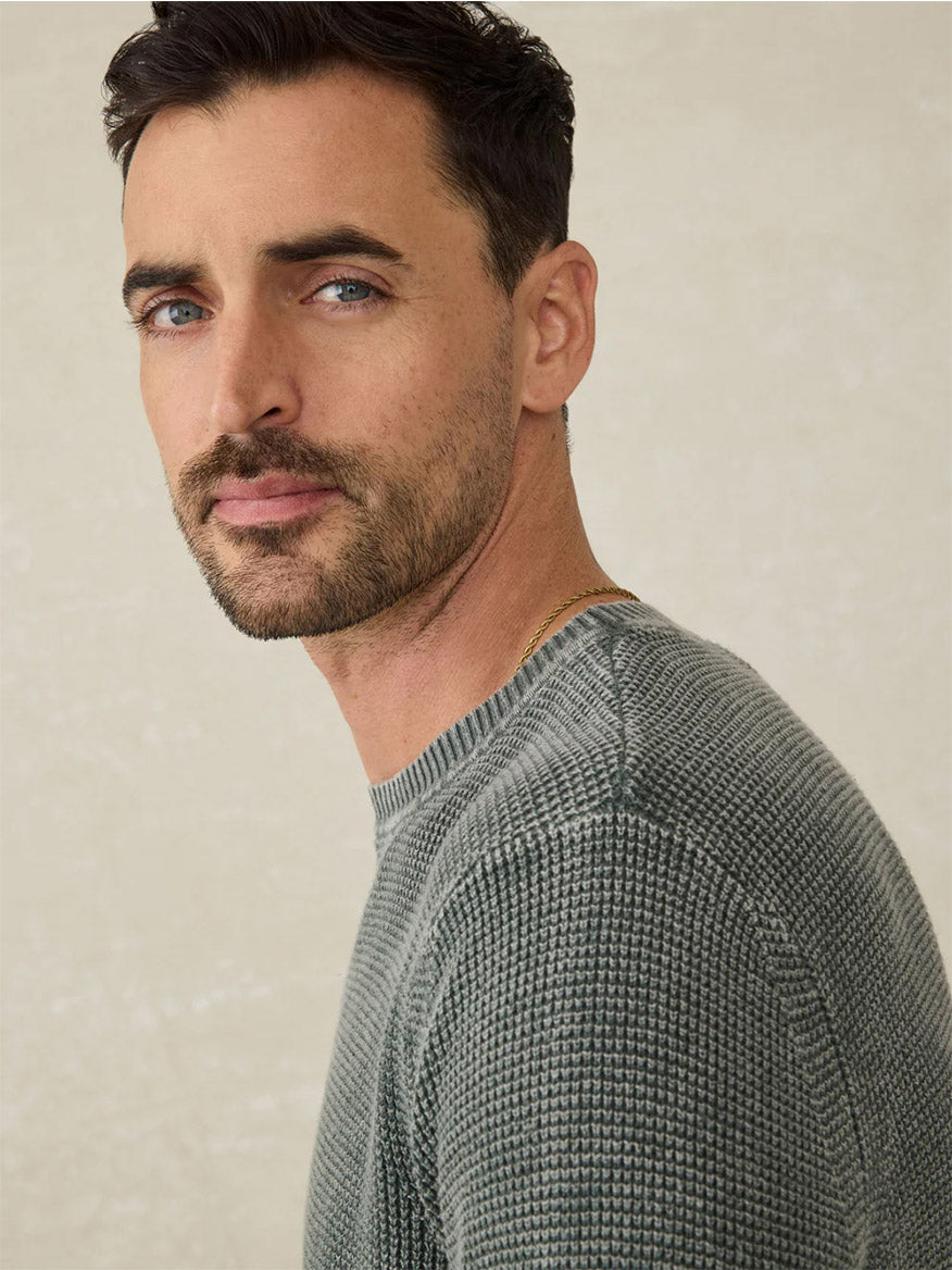 A man with short dark hair and a trimmed beard wears the Faherty Brand Sunwashed Crewneck Sweater in Mountain Olive, gazing at the camera against a light, neutral background for a vintage-inspired look.