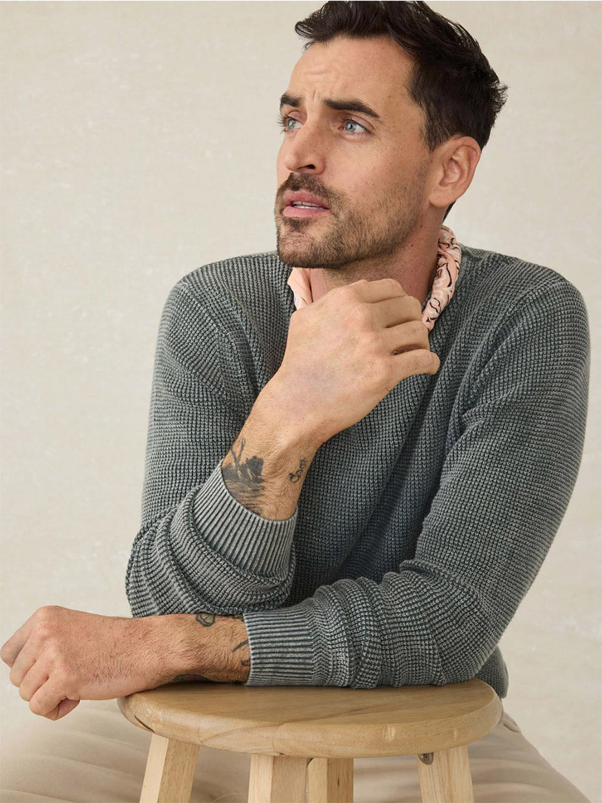 Man with short dark hair and light stubble, wearing the Faherty Brand Sunwashed Crewneck Sweater in Mountain Olive, sits on a wooden stool looking to his left with his hand near his face.
