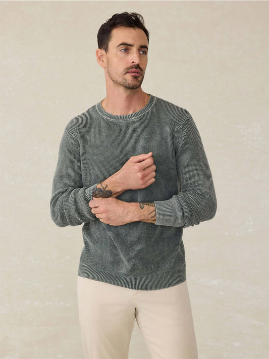 A man with short dark hair and forearm tattoos wears a Faherty Brand Sunwashed Crewneck Sweater in Mountain Olive and light beige pants, standing against a plain, light background.