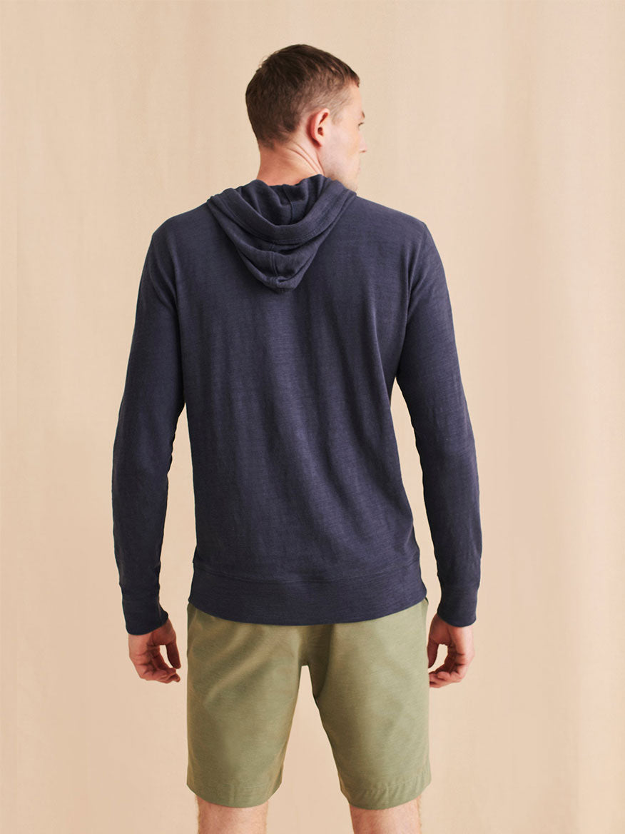 A man faces away, wearing the Faherty Brand Sunwashed Midweight Organic Hoodie in Blue Nights and olive green shorts against a plain beige background.