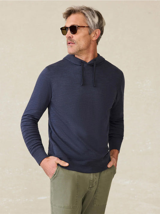 Man wearing sunglasses and the Faherty Brand Sunwashed Midweight Organic Hoodie in Blue Nights with olive green pants stands with hands in pockets against a light neutral background.