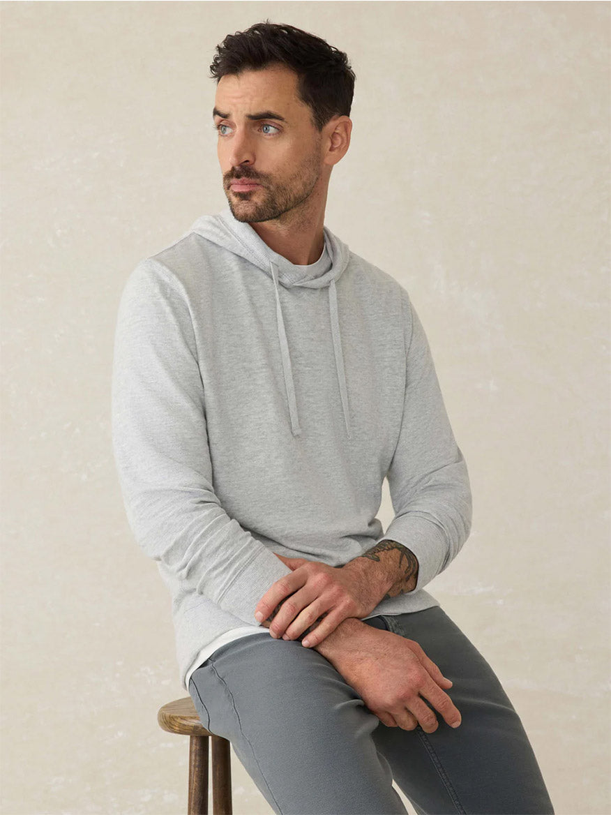 A man with short dark hair and a trimmed beard sits on a wooden stool, wearing the Faherty Brand Sunwashed Midweight Organic Hoodie in Light Grey Heather and matching gray pants, looking off to the side.