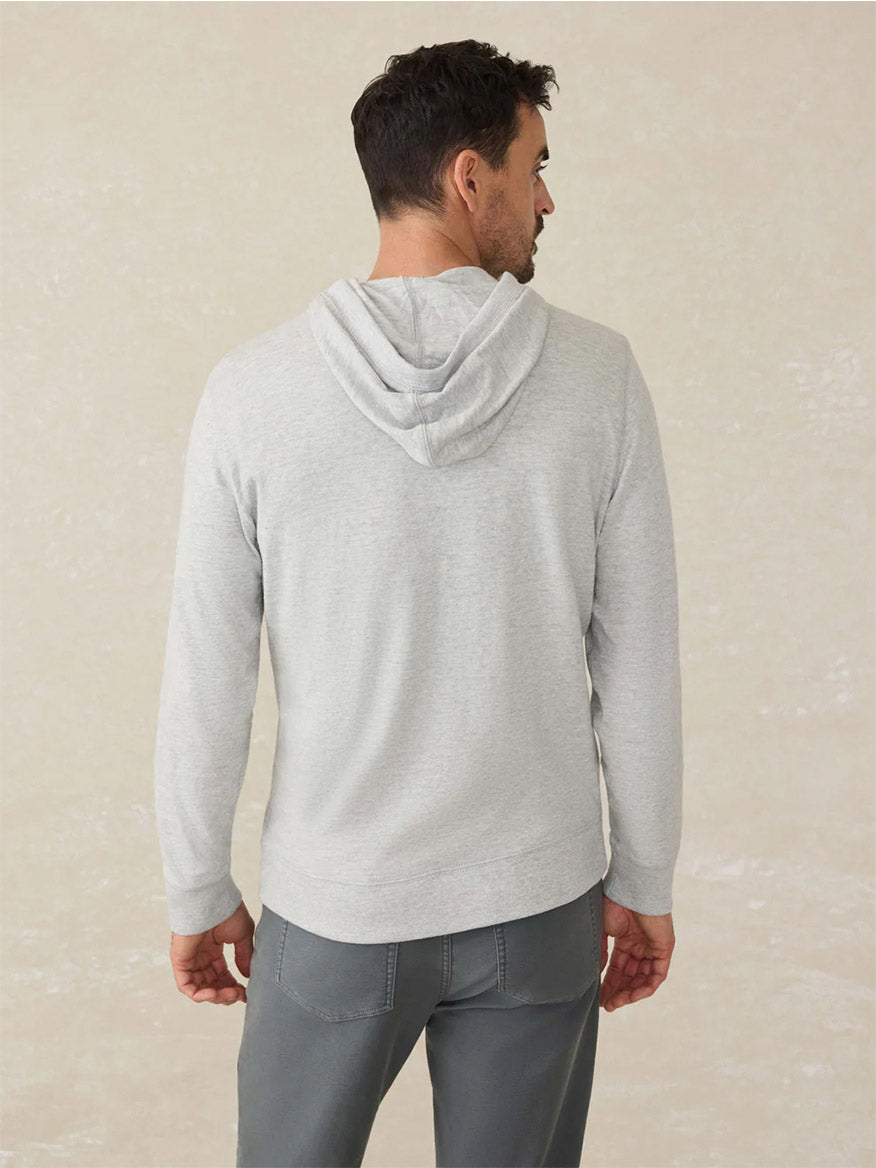 A man stands facing away, wearing a Faherty Brand Sunwashed Midweight Organic Hoodie in Light Grey Heather and gray pants made from American-grown cotton against a neutral background.